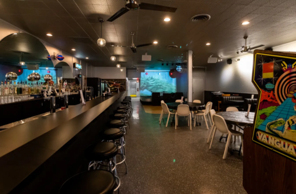 Retro-style interior of UFO Bar in Detroit featuring a long black bar with chrome stools, vintage arcade games, neon signage, and a laid-back neighborhood cocktail atmosphere.