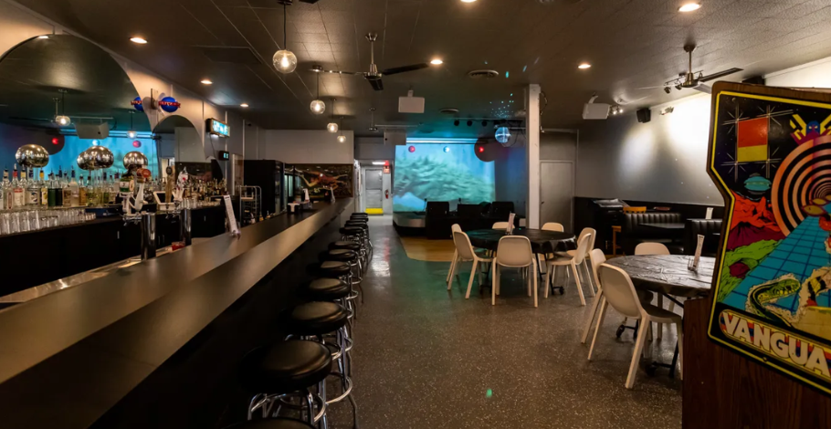 Retro-style interior of UFO Bar in Detroit featuring a long black bar with chrome stools, vintage arcade games, neon signage, and a laid-back neighborhood cocktail atmosphere.