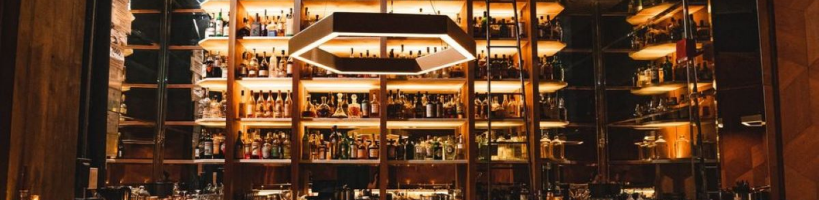 Moody interior of Bad Luck Bar in Detroit featuring a dramatic backlit wall of premium spirits, modern geometric pendant lighting, and an upscale cocktail bar with warm amber tones.