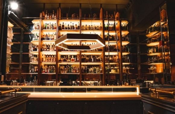 Moody interior of Bad Luck Bar in Detroit featuring a dramatic backlit wall of premium spirits, modern geometric pendant lighting, and an upscale cocktail bar with warm amber tones.