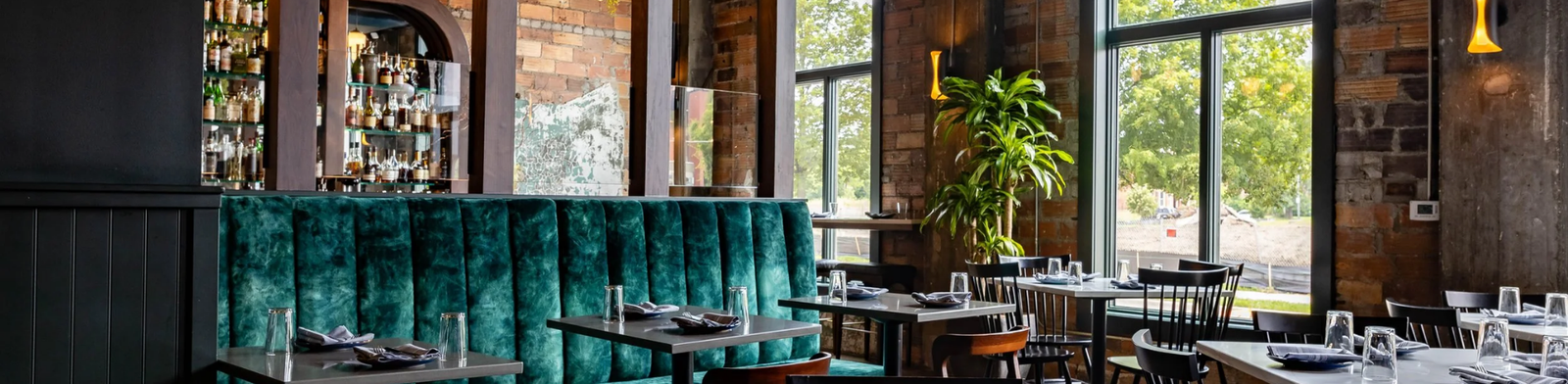 Stylish interior of Bar Pigalle in Detroit featuring emerald velvet banquette seating, warm wood arches, exposed brick walls, and a bright dining space with modern tables and natural light.