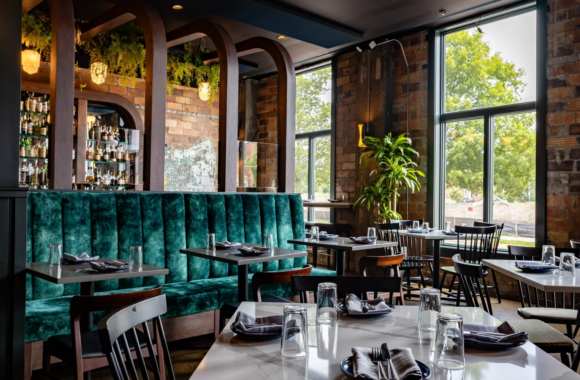 Stylish interior of Bar Pigalle in Detroit featuring emerald velvet banquette seating, warm wood arches, exposed brick walls, and a bright dining space with modern tables and natural light.