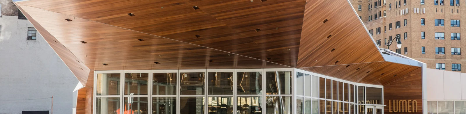 Modern exterior of Lumen Detroit restaurant in Beacon Park featuring a striking angular wood canopy, floor-to-ceiling glass walls, and outdoor patio seating, with downtown Detroit buildings in the background.