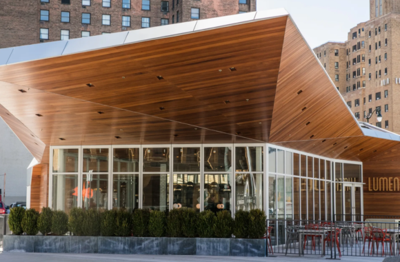 Modern exterior of Lumen Detroit restaurant in Beacon Park featuring a striking angular wood canopy, floor-to-ceiling glass walls, and outdoor patio seating, with downtown Detroit buildings in the background.