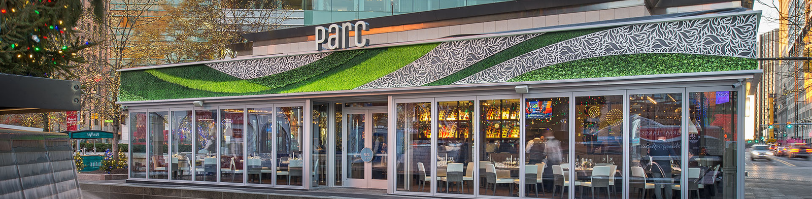 Exterior of Parc restaurant in downtown Detroit featuring a sleek glass façade, vibrant green rooftop design, and views of Campus Martius with a modern fountain in the foreground.