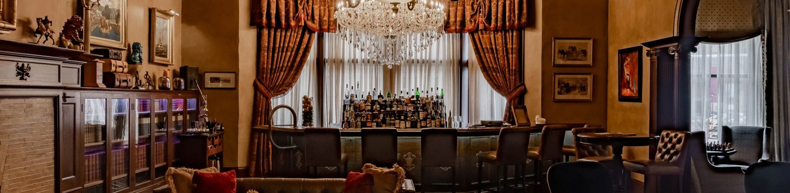 Elegant lounge interior of The Library at The Detroit Club featuring a crystal chandelier, leather seating, rich wood accents, classic drapery, and a sophisticated cocktail bar lined with spirits in a historic Detroit setting.