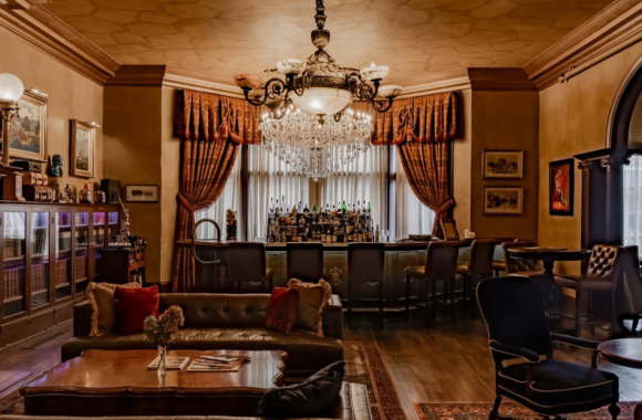 Elegant lounge interior of The Library at The Detroit Club featuring a crystal chandelier, leather seating, rich wood accents, classic drapery, and a sophisticated cocktail bar lined with spirits in a historic Detroit setting.