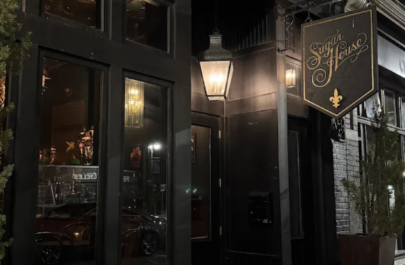 Exterior of The Sugar House in Detroit at night featuring a black façade, warm lantern lighting, and a classic hanging sign, creating an intimate and upscale cocktail bar atmosphere.