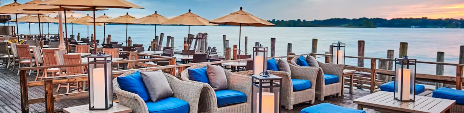 Waterfront patio at River Crab Blue Water Inn in St. Clair, Michigan featuring wicker lounge seating with blue cushions, umbrella-covered tables, and scenic views of the St. Clair River at sunset.