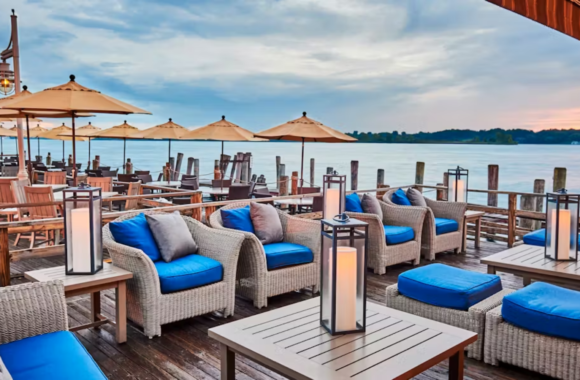Waterfront patio at River Crab Blue Water Inn in St. Clair, Michigan featuring wicker lounge seating with blue cushions, umbrella-covered tables, and scenic views of the St. Clair River at sunset.