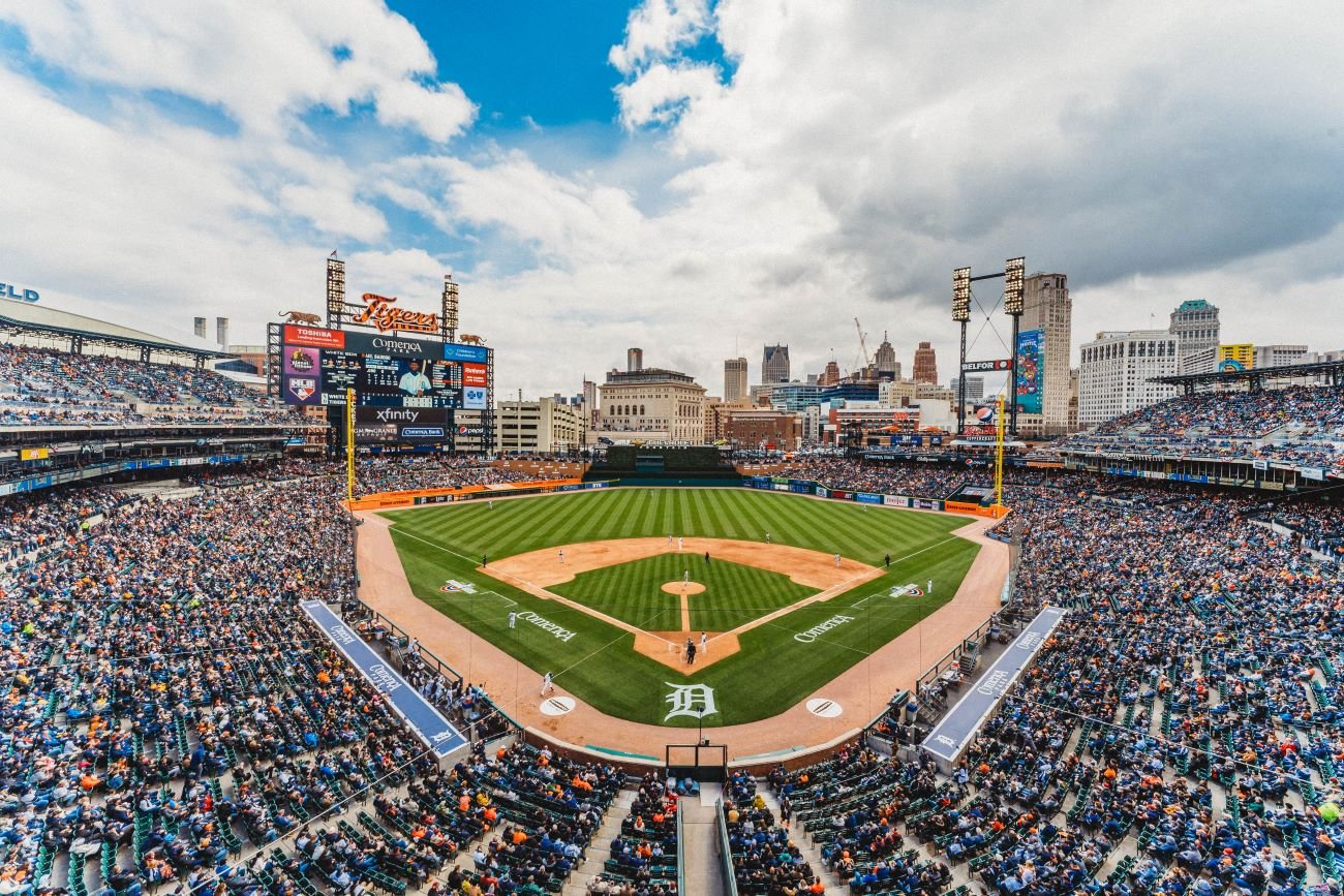 Panoramic view of a Detroit Tigers baseball game at Comerica Park on a sunny day, with players on the field and the Detroit skyline in the background.
