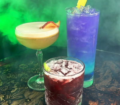 Halloween-themed cocktails at 225 Speakeasy in Atwater Brewery Detroit, featuring red, golden, and purple drinks in a smoky, moody setting.
