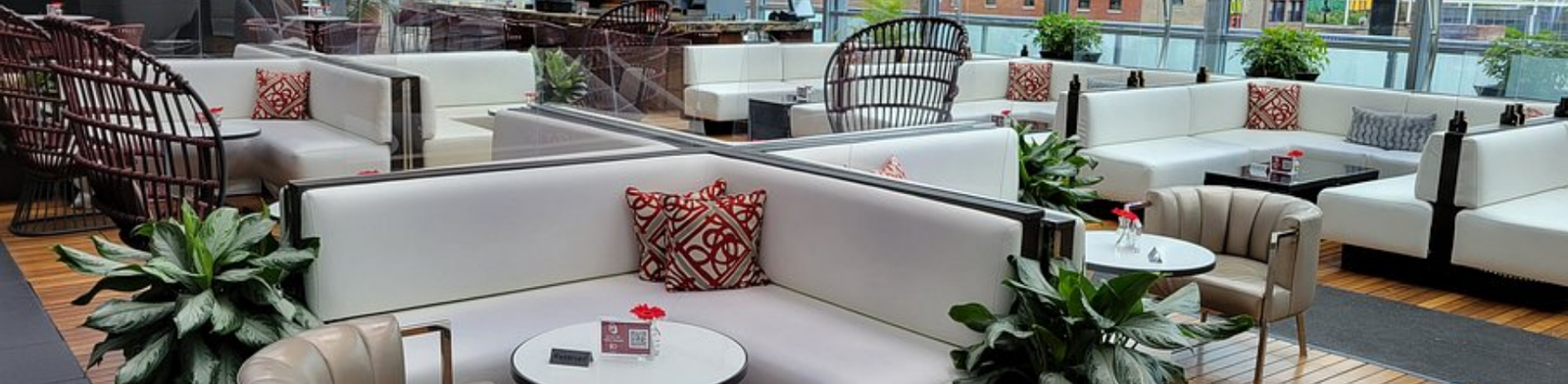 Modern rooftop lounge with white sectional seating, wicker chairs, and small cocktail tables under a glass canopy, overlooking downtown buildings with greenery and stylish terrace décor.