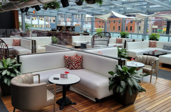 Modern rooftop lounge with white sectional seating, wicker chairs, and small cocktail tables under a glass canopy, overlooking downtown buildings with greenery and stylish terrace décor.