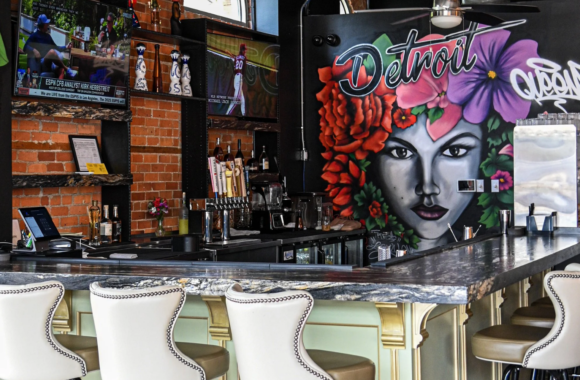 Upscale Detroit bar interior with marble counter seating and a colorful mural of a woman with flowers and “Detroit” graffiti behind the bar.