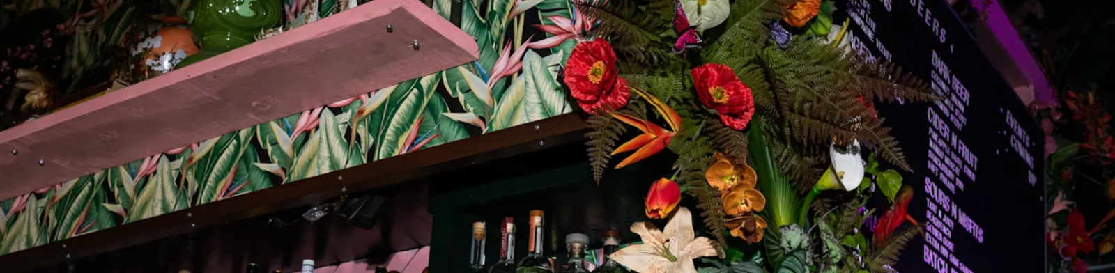 Vibrant tropical-themed interior of The Skip in Detroit featuring colorful floral décor, lush greenery, a back bar lined with spirits, and a lively cocktail menu board in a dimly lit setting.