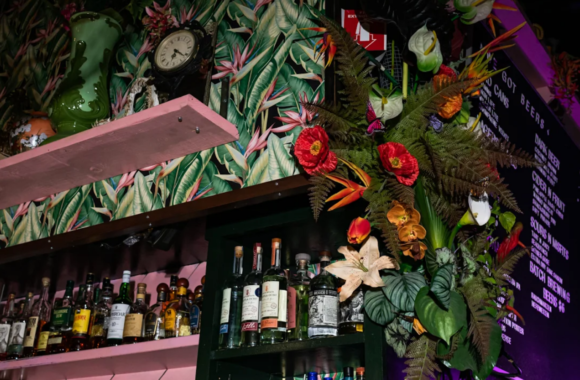 Vibrant tropical-themed interior of The Skip in Detroit featuring colorful floral décor, lush greenery, a back bar lined with spirits, and a lively cocktail menu board in a dimly lit setting.