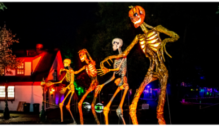 Canterbury Village Halloween Stroll featuring towering illuminated skeleton and pumpkin-headed figures glowing against a nighttime backdrop of festive lights and decorated village buildings.