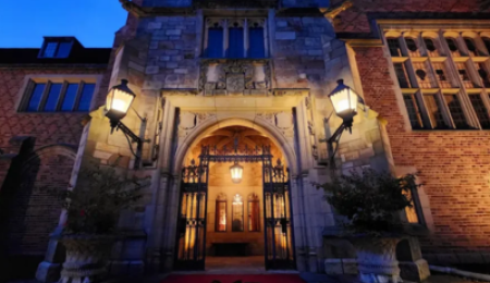 Meadow Brook Hall entrance at dusk, featuring elegant Gothic-style architecture, stone archway, wrought-iron gates, and warm golden lighting illuminating the historic mansion.