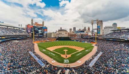 Panoramic view of a Detroit Tigers baseball game at Comerica Park on a sunny day, with players on the field and the Detroit skyline in the background.